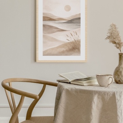 Cadeira de madeira clara ao lado de uma mesa com toalha bege, livro aberto, caneca, vaso com planta seca e quadro com pintura em tons sépia de paisagem abstrata na parede.