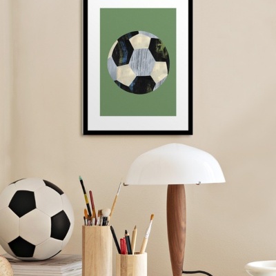 Quadro com bola de futebol estilizada verde pendurado numa parede bege acima de mesa com bola de futebol real, porta-lápis, lâmpada e material de escritório
