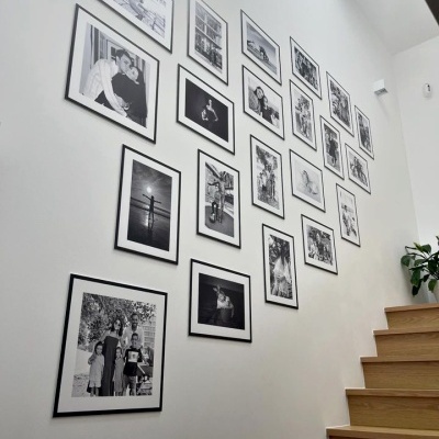 Parede branca com vários quadros em molduras pretas com fotos a preto e branco organizados diagonalmente junto a escada de madeira clara.