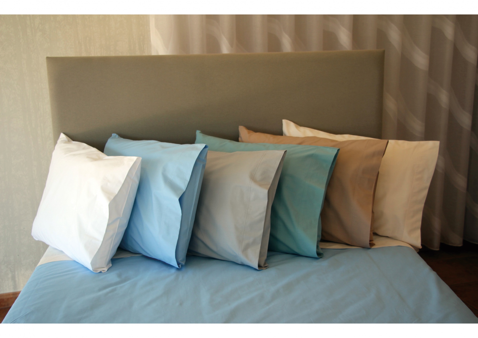 Almofadas de cama coloridas alinhadas num colchão com lençol azul e cabeceira cinza