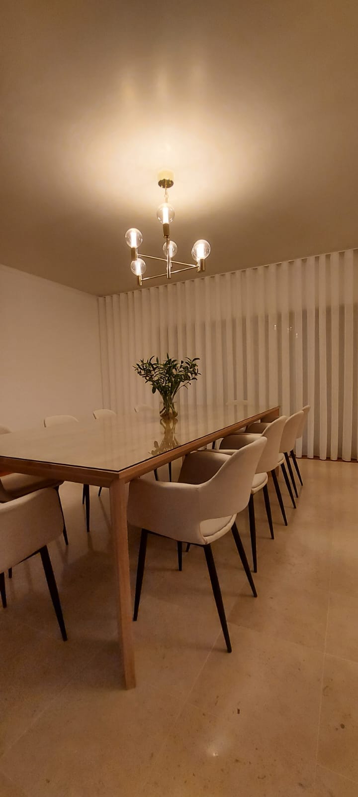 Mesa de jantar de madeira com tampo de vidro e cadeiras estofadas bege numa sala com cortinas brancas e candeeiro moderno de teto