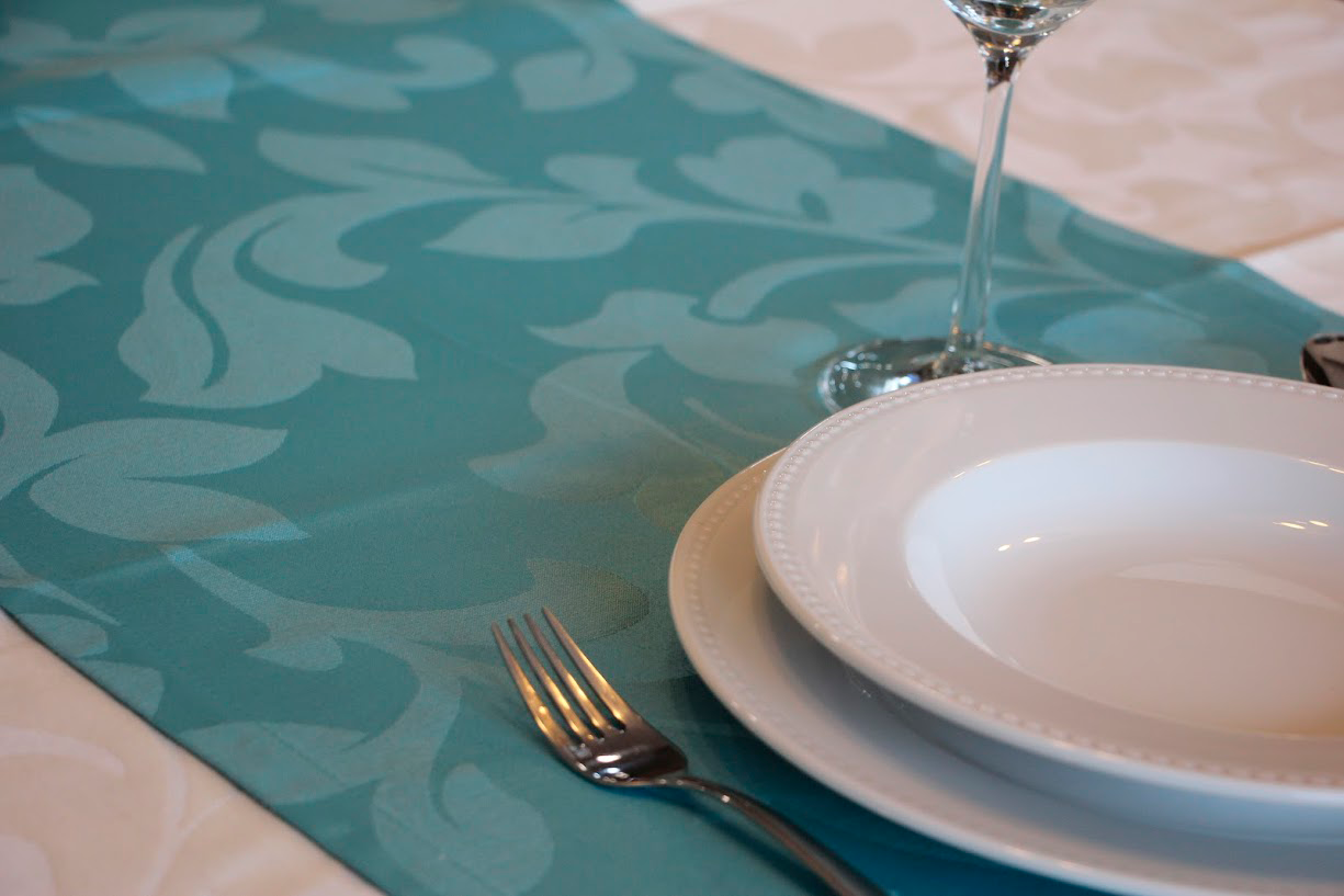 Mesa posta com toalha branca e caminho de mesa azul-claro, pratos brancos, garfo e copo