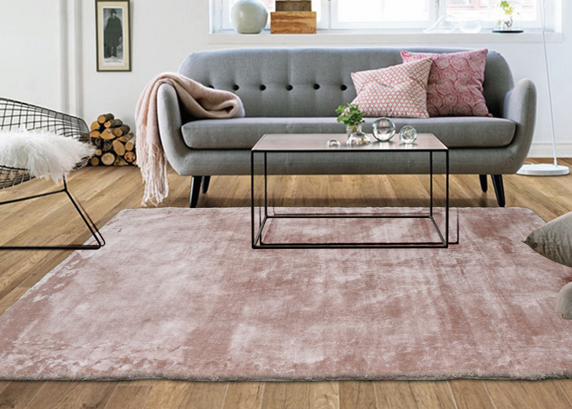 Sala de estar com sofá cinza, almofadas rosas, manta, mesa de centro de vidro, tapete rosa claro e cadeira metálica com assento branco felpudo