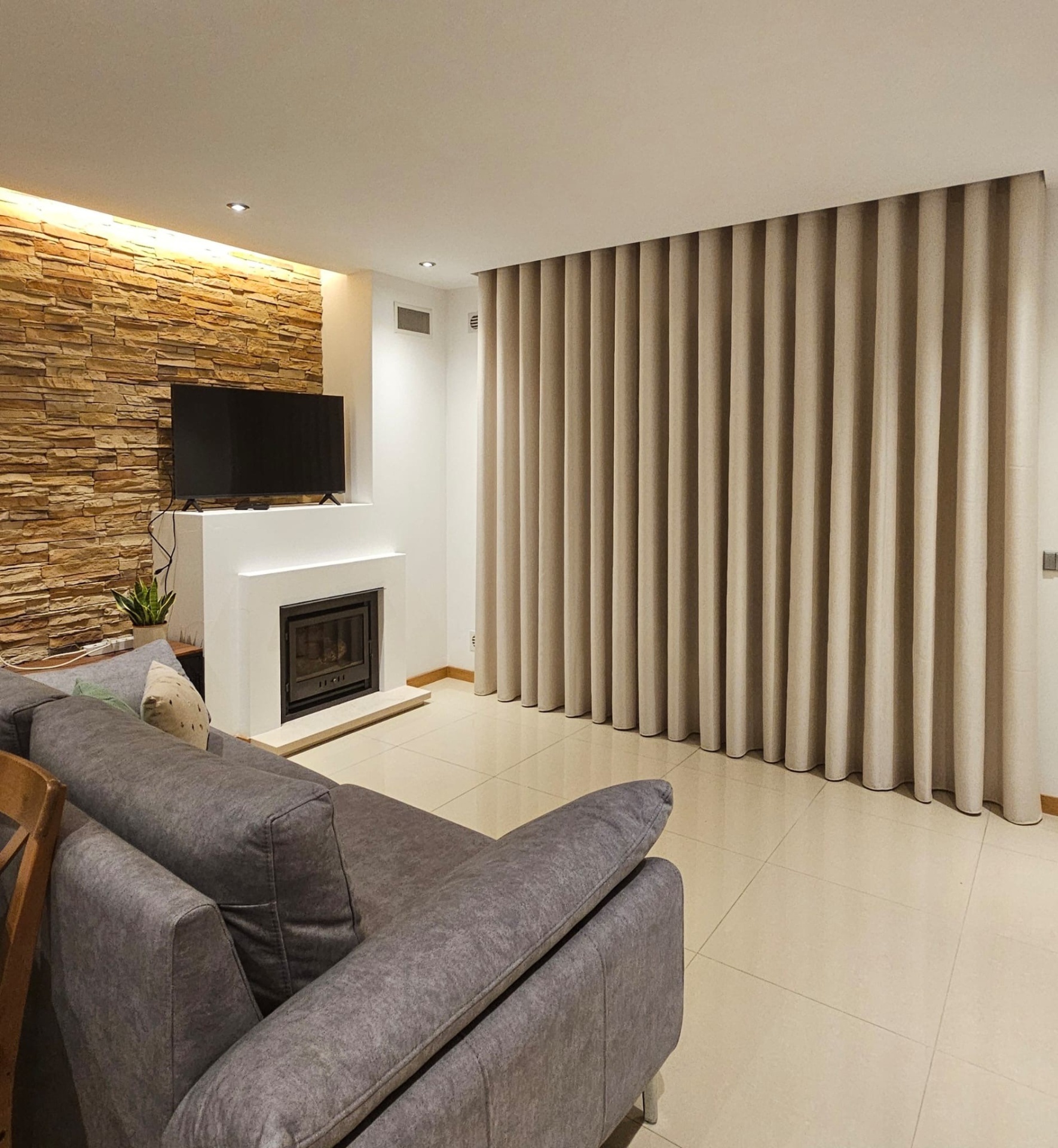 Sala de estar com sofá cinza, cortinas bege e parede decorativa de pedra
