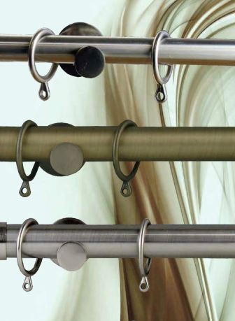 Três varões de cortinas metálicos em diferentes tons com argolas e suportes, fundo abstrato desfocado em castanho e creme.