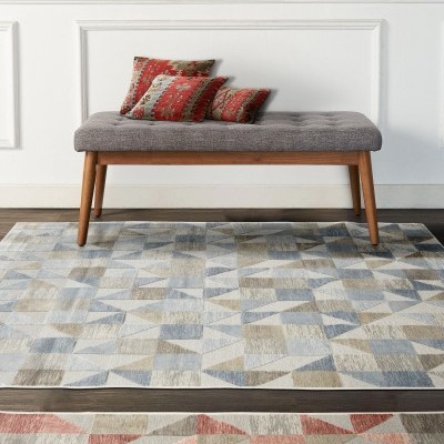 Banco cinza de botão acolchoado com pernas de madeira e almofadas vermelhas em tapete geométrico azul e bege