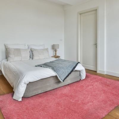 Quarto com cama branca, almofadas bege, manta cinzenta e tapete rosa