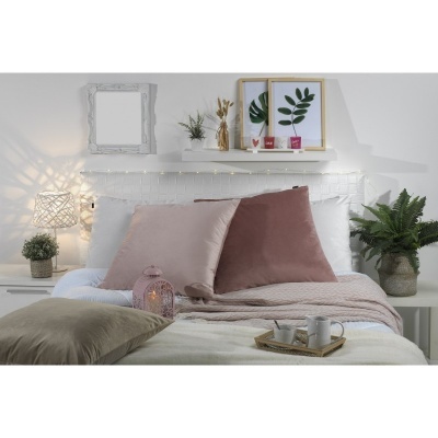 Cama com almofadas rosas, manta bege, bandeja com canecas, plantas e quadros na parede branca