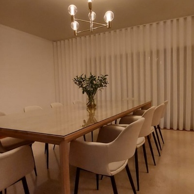 Mesa de jantar de madeira com tampo de vidro e cadeiras estofadas bege numa sala com cortinas brancas e candeeiro moderno de teto