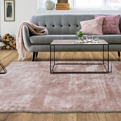 Sala de estar com sofá cinza, almofadas rosas, manta, mesa de centro de vidro, tapete rosa claro e cadeira metálica com assento branco felpudo