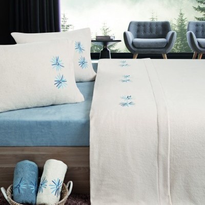 Roupa de cama branca com bordados azuis sobre lençol azul numa cama, com poltronas cinza ao fundo