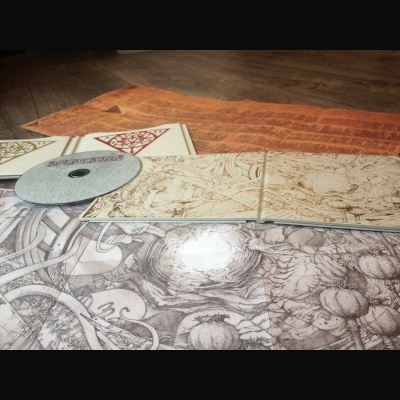 Folhas e newsletters artísticas em tons sépia e cinza junto a um CD prateado sobre madeira e tapete laranja