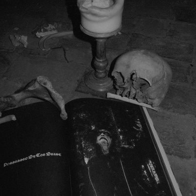 Caveira, vela derretida em castiçal, osso e página escura de revista com texto 'Doomed By The Dead' no chão de pedra.