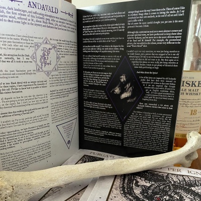 Livro aberto com texto, osso sobreposto, garrafa de uísque e livros empilhados em mesa de madeira clara