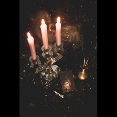 Castiçal com três velas acesas, copo metálico com varetas, livro escuro e espinho sobre superfície preta