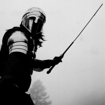 Pessoa com armadura metálica e capacete a segurar uma espada longa em ambiente nebuloso