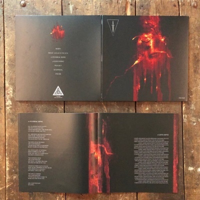 Capa aberta de CD ou vinil com arte vermelha abstrata e texto em branco sobre fundo preto, acompanhado de livreto com letras e texto em inglês, sobre chão de madeira.