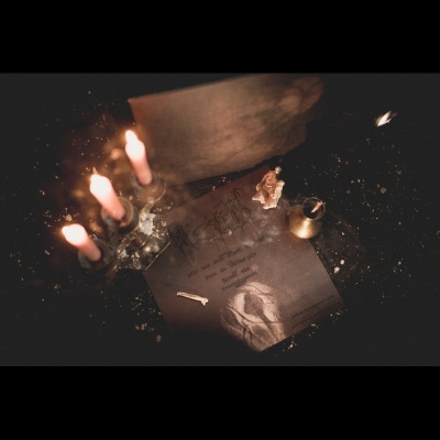 Quatro velas acesas num castiçal, papel queimado, frasco dourado e pano branco sobre superfície escura