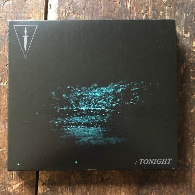 Caixa preta com imagem azul turquesa abstrata, texto TONIGHT e símbolo de adaga, sobre mesa de madeira.