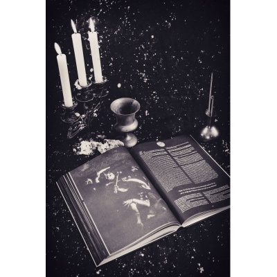 Livro aberto com ilustração e texto, castiçal com velas acesas, cálice metálico e castiçal com vela apagada sobre superfície escura