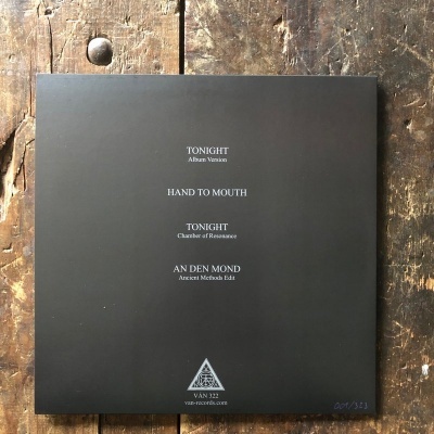 Capa de disco vinil preta com títulos das faixas e logotipo triangular sobre fundo de madeira