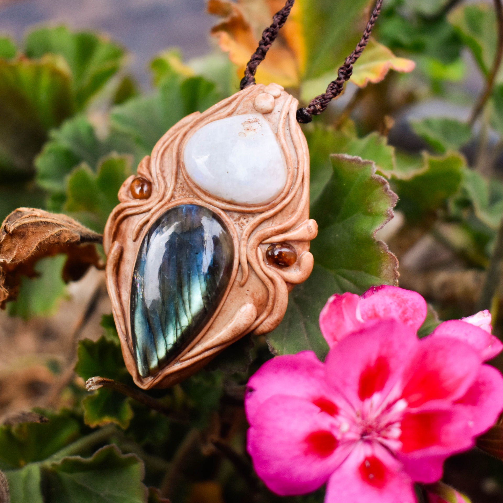 Amuleto Dharma com Pedra da Lua, Turmalinas e Labradorite Pingente artesanal com pedras brancas e azul esverdeadas em ambiente natural com flor rosa