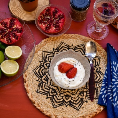 Pequeno-almoço com iogurte, frutas, colher e guardanapo sobre base decorativa redonda.