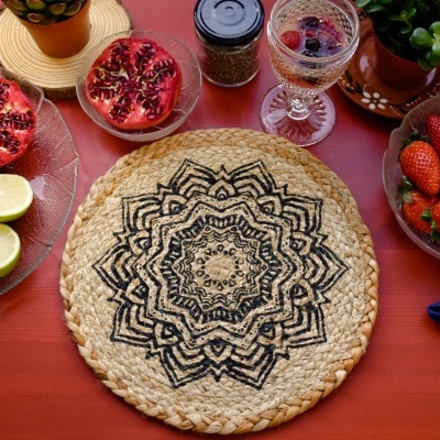 Base de mesa redonda em fibras naturais com padrão preto, rodeada de frutas e bebida numa mesa vermelha