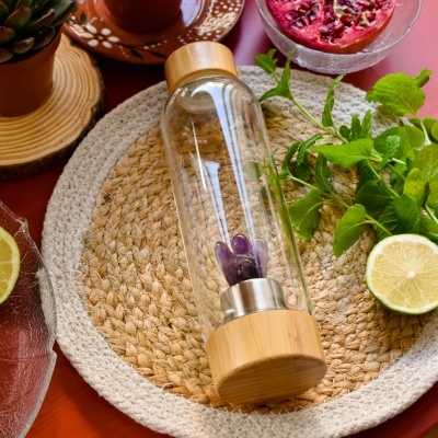 Garrafa de vidro com tampas de madeira e pedras roxas no interior sobre tapete redondo de fibras naturais