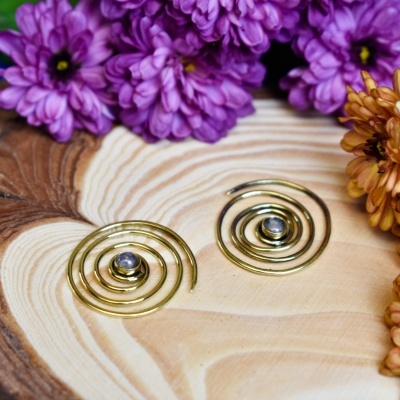 Brincos espirais dourados com pedra central sobre madeira e flores ao redor