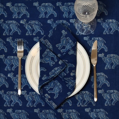 Mesa posta com toalha e guardanapo azul com padrão de elefantes, prato branco com borda dourada, talheres metálicos e copo de cristal