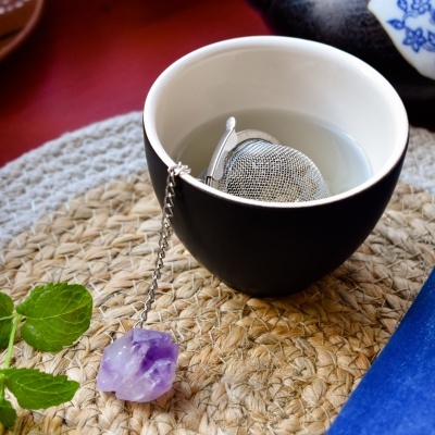 Infusor de chá metálico numa chávena preta com pedra roxa pendurada, sobre esteira de palha.