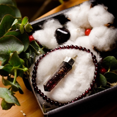 Conjunto de joalharia com pulseira de contas vermelhas, pendente de pedra castanha e preta, pedra preta, em caixa preta com algodão e folhas verdes