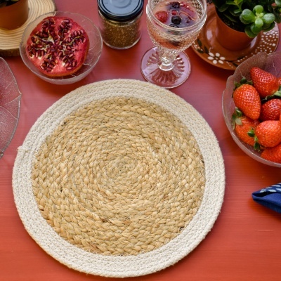 Manta redonda de fibras naturais bege e branca sobre mesa vermelha com frutas e planta