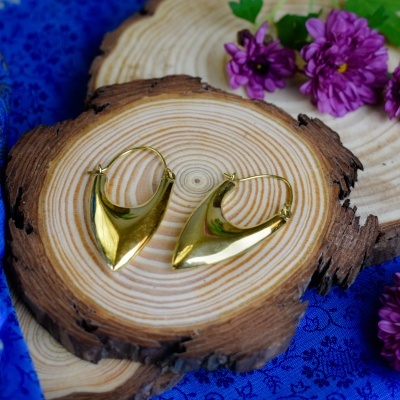 Brincos dourados geométricos sobre base de madeira com flores roxas e tecido azul