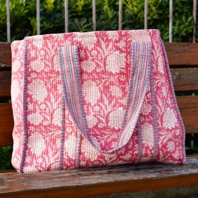 Mala de tecido rosada com padrão floral branco e alças riscas sobre banco de madeira