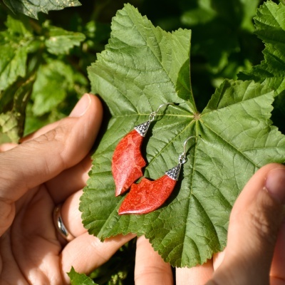 Brincos pendentes vermelhos com topo metálico prateado sobre folha verde segura por mãos