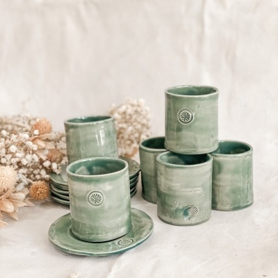 Copos e pratos verdes em cerâmica com selo de folha, sobre superfície branca com flores secas ao lado