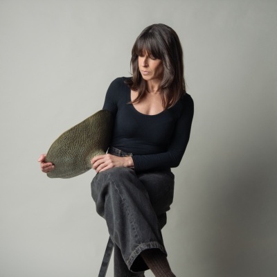 Mulher sentada num banco alto a segurar prato decorativo verde oval com padrão texturizado