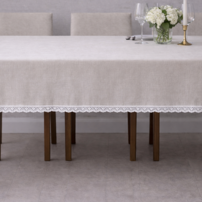 Toalha de mesa bege com borda de renda branca sobre mesa de madeira, com vaso de flores brancas, castiçal e copos ao fundo