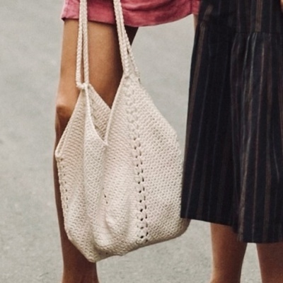 Duas mulheres com sandálias rasas castanhas, uma com vestido rosa e bolsa de malha branca, outra com saia preta às riscas