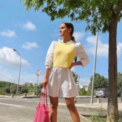 Mulher com vestido branco, colete amarelo, tênis branco e bolsa rosa na rua