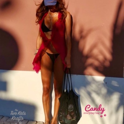 Mulher de biquíni preto com colete vermelho e chapéu azul DEUS segurando mala preta tipo rede em calçada junto a parede castanha