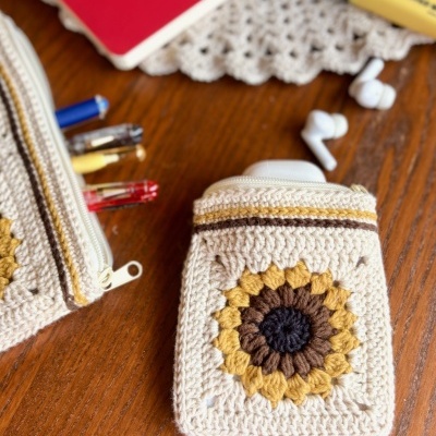 Duas bolsas de croché bege com padrões circulares coloridos, uma com canetas e outra com caixa branca, em mesa de madeira com toalha de renda.