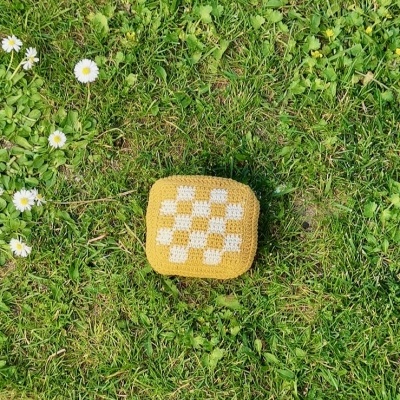 Almofada quadrada tricotada amarela com padrão xadrez branco sobre relva verde com flores brancas