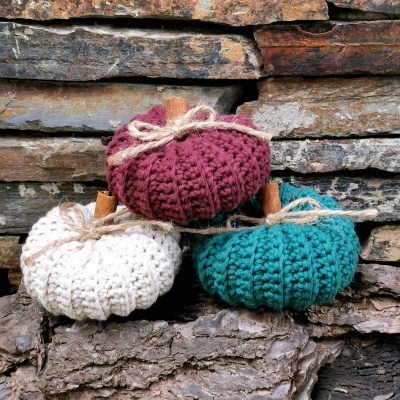 Três abóboras decorativas de crochet em vermelho, branco e verde com pauzinhos e cordões, sobre troncos e pedras.