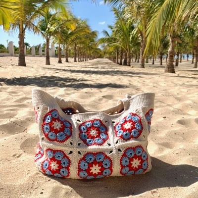 Mala de croché colorida na areia de praia com palmeiras ao fundo.