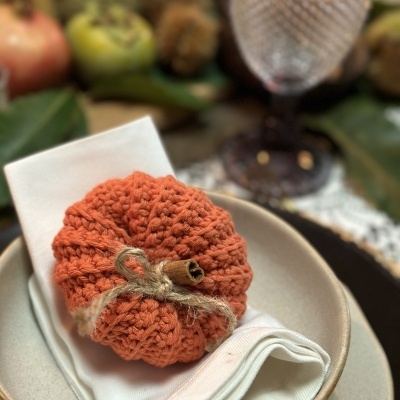 Guardanapo branco com abóbora de tecido tricotado laranja numa tigela beige, copo de vidro e decoração de folhas e frutas ao fundo