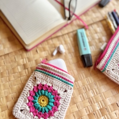 Estojo de malha colorido com estojo branco e acessórios sobre mesa de rattan.