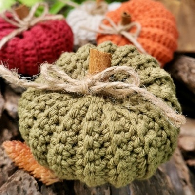 Abóboras decorativas de tricô verde, vermelho e laranja com paus de canela e laços de corda rústica sobre madeira.